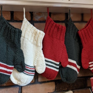 May include: Five knitted Christmas stockings in various colours hang from a mantelpiece. The stockings are dark green, cream, and red, with white and red stripes near the cuff. The stockings are handmade and have a cosy, festive appearance.