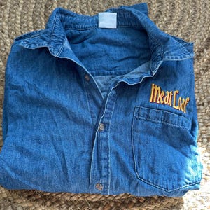 May include: A blue denim button-up shirt with a pocket on the left chest. The shirt has the embroidered text "Meat Loaf" in yellow and orange thread on the right chest. The shirt is folded on a woven surface.