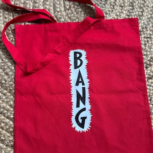 May include: A red tote bag with long straps. The word "BANG" is printed vertically in black letters on a white, jagged-edged rectangle. The bag is made of a sturdy fabric.
