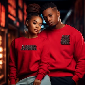 Peut inclure: Deux personnes portant des sweat-shirts rouges assortis. Chaque sweat-shirt porte le mot "AMOR" dans une police sombre et scintillante. L'arrière-plan est flou, avec des lumières aux tons chauds.