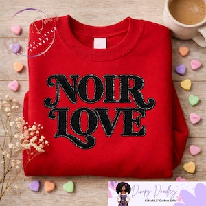 May include: A red sweatshirt with the words "NOIR LOVE" embroidered in black, glittery thread. The sweatshirt is displayed on a wooden surface, with heart-shaped candies and a cup of coffee nearby. The sweatshirt is a piece of apparel.