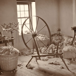 Könnte beinhalten: Ein Spinnrad mit einem großen Holzrad und einer Spindel, umgeben von einem Haufen weißer Wolle. Im Vordergrund befindet sich ein Korb mit Wolle und ein Holztisch mit einem Spinnrad.