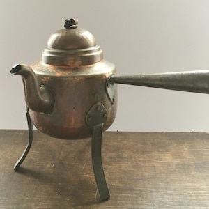 Puede incluir: Una tetera de cobre con tapa abovedada y un largo pico. La tetera se apoya en tres patas de hierro.