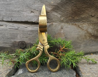 May include: A pair of vintage-style brass scissors featuring a polished metallic finish, two circular finger loops, and a distinct cylindrical bolster located near the pivot point, designed with a classic aesthetic and a sturdy metal construction.
