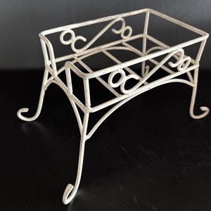 May include: A rectangular off-white metal stand featuring a wire frame construction with decorative scrollwork details along the sides. The stand is supported by four slender, curved legs that extend outward, providing a stable base for the open top.