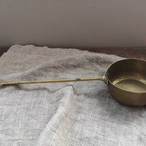 May include: A brass ladle with a long handle and a square-shaped end. The ladle is sitting on a light brown linen fabric.