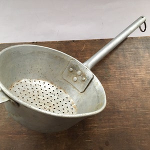 May include: A silver aluminum strainer with a long handle and a perforated bottom. The strainer has a small flat handle on the side and a ring on the end of the handle.