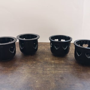 以下が含まれることがあります： ハート型の切り抜きのある黒いセラミック製のキャンドルホルダーが4つ。ホルダーは木製の表面に一列に並んでいます。