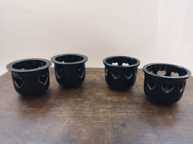 以下が含まれることがあります： ハート型の切り抜きのある黒いセラミック製のキャンドルホルダーが4つ。ホルダーは木製の表面に一列に並んでいます。