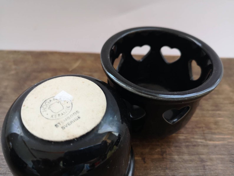 以下が含まれることがあります： ハート型の切り抜きのある黒いセラミック製のキャンドルホルダー2つ。1つのホルダーの底には、「Svenska Keramikk, Stengods, Sverige」というテキストが書かれた白いラベルがあります。