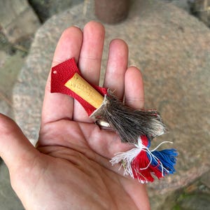 Può includere: Un piccolo ciondolo decorativo con un pezzo centrale in legno chiaro inciso con il testo 'WISTEN'. Il legno è rivestito in pelle rossa testurizzata e pelliccia marrone, rifinito con una nappa multicolore nei toni del rosso, bianco e blu.