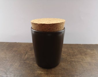 May include: A dark brown ceramic jar with a cork lid. The jar is sitting on a wooden surface.