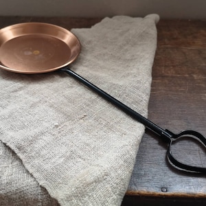 May include: A copper pan with a black metal handle. The pan is round and has a slightly raised edge. The handle is long and has a loop at the end.