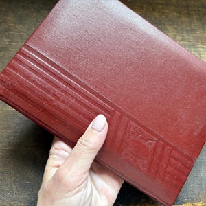 May include: A burgundy book with a textured cover. The cover has parallel lines and a geometric design. The book is held in a hand.