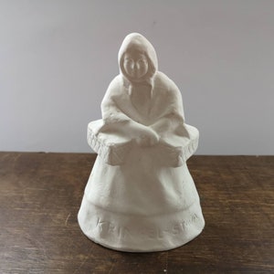Può includere: Figurina in ceramica bianca di una donna che indossa un mantello con cappuccio e tiene un cestino. La figurina è incisa con il testo "KRIST STAVA".