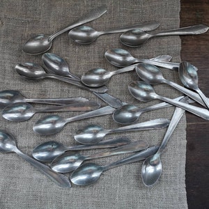 May include: A pile of silver spoons on a beige linen surface. The spoons are arranged in a random pattern, with some overlapping. The spoons are all the same size and shape, and they have a shiny, polished finish.