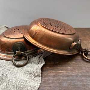 May include: Two round copper strainers with perforated surfaces and attached metal rings for hanging. The strainers feature a warm metallic finish and a shallow bowl shape, with one positioned slightly behind the other to show the design.