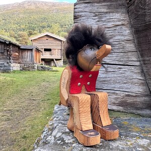 Pode incluir: Uma figura de troll de madeira com um nariz grande, cabelo preto e um colete vermelho com botões prateados. O troll está sentado numa parede de pedra, com edifícios de madeira e uma montanha ao fundo.