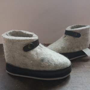Könnte beinhalten: Ein Paar graue Filzpantoffeln mit schwarzem Lederbesatz und einem Knopfverschluss. Die Pantoffeln haben eine schwarze Gummisohle.