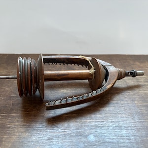 Könnte beinhalten: Ein hölzernes Spinnrad mit Spindel und Kreisel. Das Rad ist aus dunklem Holz und hat einen Metallhaken an der Seite. Der Kreisel ist aus Metall und hat eine Reihe von Rillen.