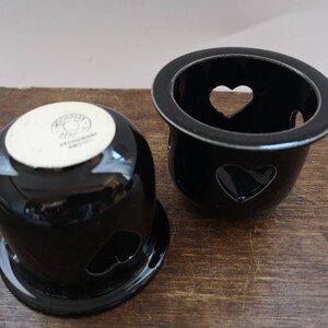 以下が含まれることがあります： ハート型の切り抜きのある黒いセラミック製のキャンドルホルダー2つ。下のホルダーには「STEINHARDT GERMANY」という文字が書かれています。