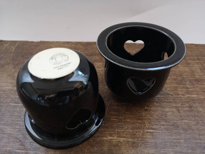 以下が含まれることがあります： ハート型の切り抜きのある黒いセラミック製のキャンドルホルダー2つ。下のホルダーには「STEINHARDT GERMANY」という文字が書かれています。