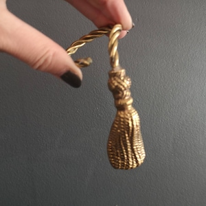 May include: A gold-colored metal tassel with a twisted rope design. The tassel is attached to a ring.