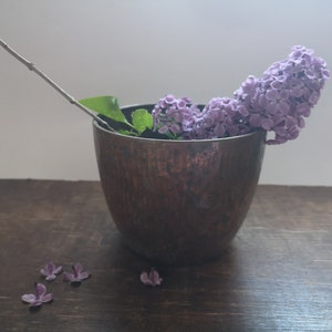 Könnte beinhalten: Eine Kupferschale gefüllt mit lila Fliederblüten und einem einzelnen Zweig. Die Schale steht auf einer dunklen Holzoberfläche.