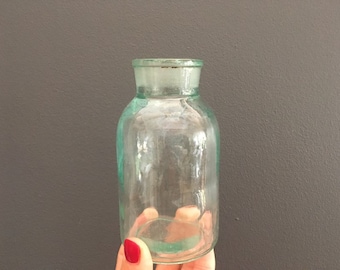 Puede incluir: Una botella de vidrio transparente con un tono verde y una forma redondeada. La botella tiene una superficie lisa y ligeramente texturizada.