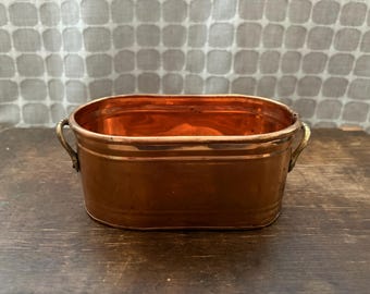 May include: An oval-shaped copper basin featuring a polished metallic finish and two small, curved brass handles attached to the sides. The container has a smooth, rounded rim and a deep, open interior, showcasing the warm, reflective orange-brown tones.