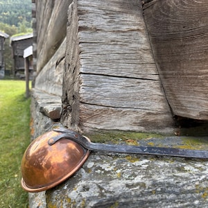 Op de afbeelding: Een koperen kookpot met een lange, donkere metalen handgreep rust op een stenen muur. De pot heeft een afgeronde komvorm en staat tegen een achtergrond van verweerde houten structuren en groen gras.