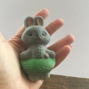 Op de afbeelding: Een grijs keramiek konijnenbeeldje met een groen geverfde broek. Het konijn heeft een lange neus, kleine zwarte ogen en een kleine roze neus.