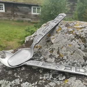 May include: Two silver spoons with ornate designs, one with a spoon bowl and the other with a fork-like end, resting on a gray rock with green moss.