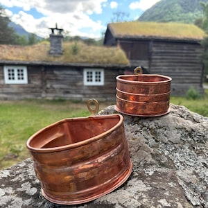 May include: Two polished copper containers with a rounded front and a flat back, each featuring a small metal loop at the top center for hanging. The containers have a smooth, reflective surface with visible horizontal bands and a warm metallic tone.