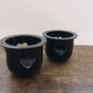 以下が含まれることがあります： ハート型の切り抜きのある黒いセラミック製のキャンドルホルダー2つ。ホルダーは木製の表面の上に置かれています。