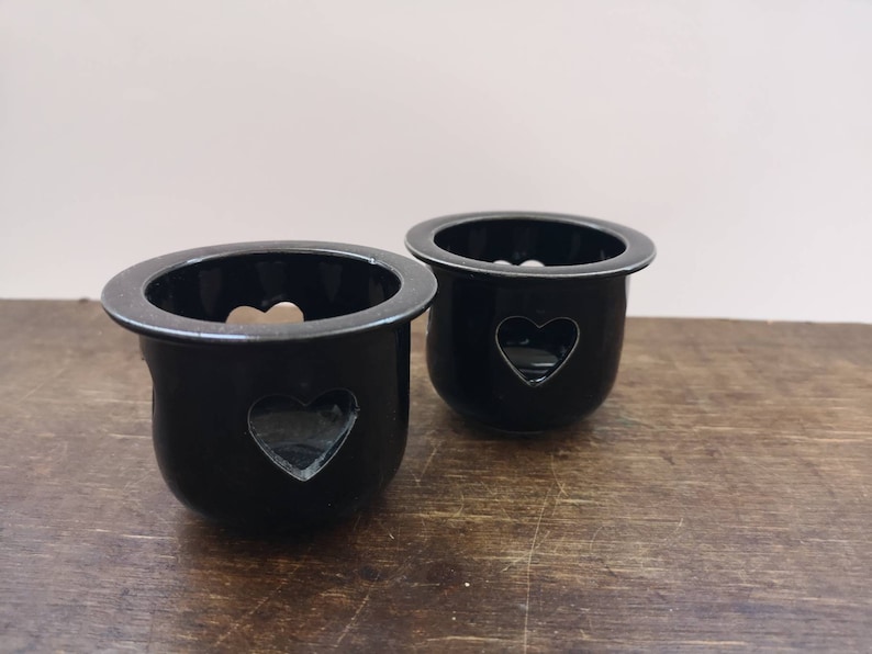以下が含まれることがあります： ハート型の切り抜きのある黒いセラミック製のキャンドルホルダー2つ。ホルダーは木製の表面の上に置かれています。