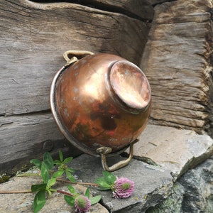 May include: A vintage copper bowl with brass handles. The bowl is slightly tarnished and has a rustic appearance. It is sitting on a stone surface with green plants and pink flowers.