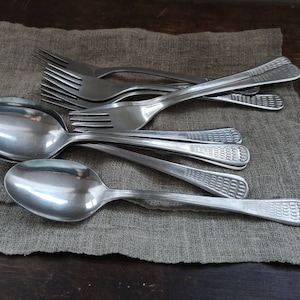 Può includere: Un set di cucchiai e forchette d'argento su un tovagliolo di lino beige. I cucchiai hanno un motivo decorativo sul manico.