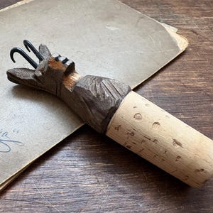 Può includere: Un tappo per bottiglia in legno fatto a mano con un design a testa di cervo intagliata. Il tappo ha una base in sughero e due ganci in metallo. La testa di cervo è marrone con accenti neri.