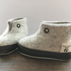 Könnte beinhalten: Ein Paar graue Filzpantoffeln mit schwarzen Gummisohlen. Die Pantoffeln haben einen Knopf oben und ein Etikett mit der Aufschrift "Norsk Husflid Engros".