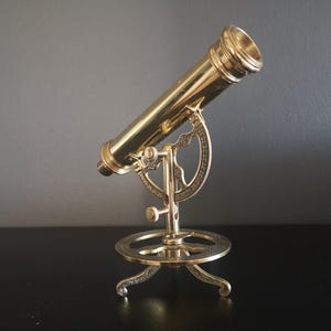 Könnte beinhalten: Ein vergoldetes Messingteleskop auf einem Stativ. Das Teleskop ist nach oben gerichtet und das Stativ hat eine runde Basis mit drei Beinen.