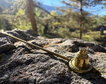 May include: A brass smoking pipe with a bell-shaped bowl and long stem rests on a textured rock. The pipe is golden in color and appears to be made of metal. The background is blurred, showing trees and a mountain.
