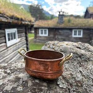 Puede incluir: Una olla ovalada de cobre con dos asas de latón en los lados opuestos. La olla presenta un acabado metálico liso con una base ligeramente redondeada y una cresta horizontal distintiva alrededor del cuerpo exterior.