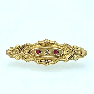 May include: A gold-toned bar pin with an oval center featuring two red gemstones and a central starburst design. The pin has ornate detailing and a decorative, elongated shape, suitable for a brooch or other jewelry.