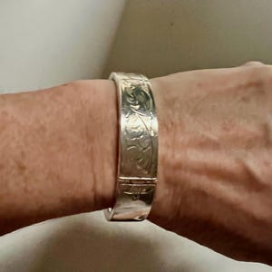 May include: A silver-coloured hinged bangle bracelet with an engraved floral design. The bracelet is worn on a wrist and has a clasp closure. The metal has a polished finish, reflecting light.