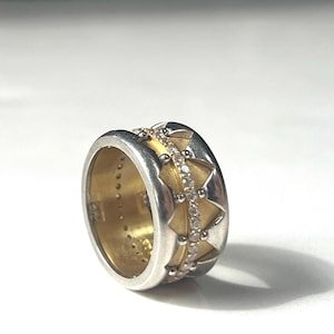 May include: A silver and gold ring with a geometric design. The ring features a band of small diamonds and a pattern of gold triangles. The ring has a wide band and a polished finish.