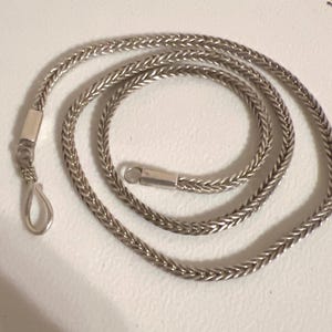May include: A silver-coloured chain necklace with a woven design. The necklace is laid out in a spiral pattern, with a hook clasp on one end and a small bar clasp on the other. The chain is made of small, interlocking links.