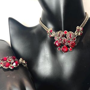 May include: A silver-toned necklace and bracelet set. The necklace features a floral design with large, red, faceted gemstones. The bracelet mirrors the design. Both pieces are displayed on a black mannequin.
