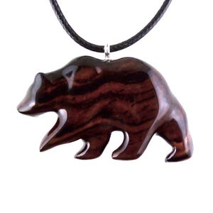 A wooden bear pendant necklace. The pendant is carved from dark reddish-brown wood with a visible grain pattern. The bear is depicted in a walking pose and is attached to a black cord necklace with a silver-colored clasp.