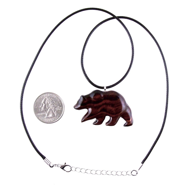 A dark reddish-brown wooden bear pendant necklace. The bear pendant is carved in a walking pose and hangs from a black cord necklace. A quarter is next to the pendant for size comparison.